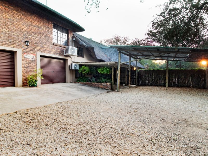 Front exterior view featuring a brick facade, double garage with roller doors, thatched roof section, and a gravel driveway with a covered carport area.