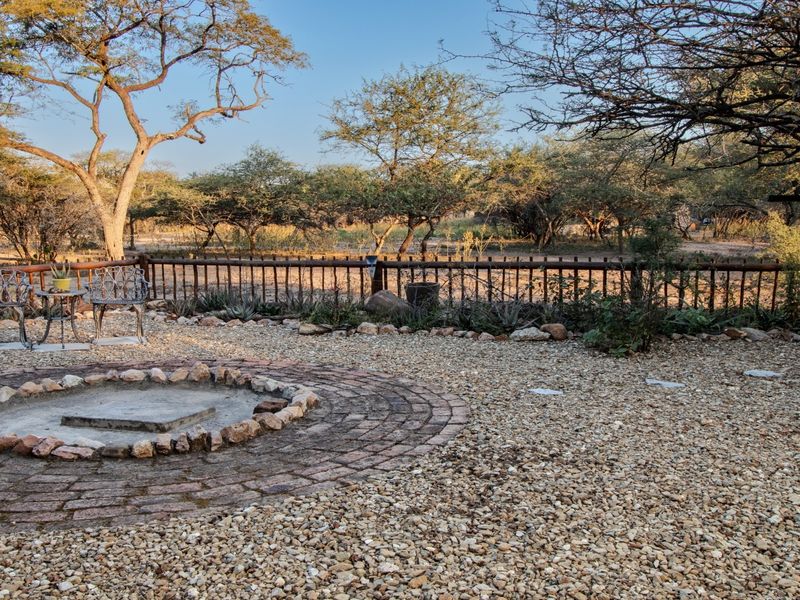 A natural outdoor garden area featuring a circular stone fire pit, gravel ground cover, and rustic timber fencing set against a backdrop of mature trees.
