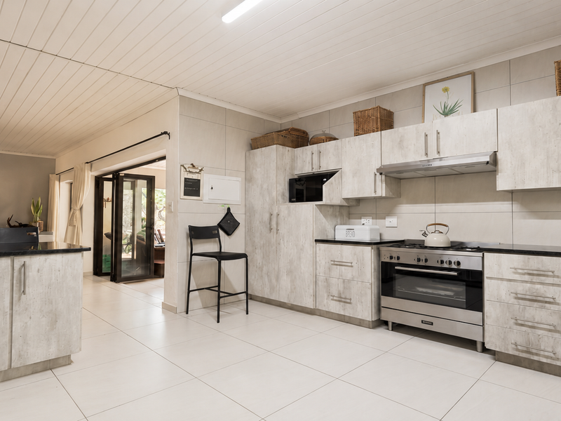 Modern kitchen featuring light wood-grain cabinetry, stainless steel gas stove with extractor fan, built-in microwave, and tiled flooring, leading to an adjacent living area.
