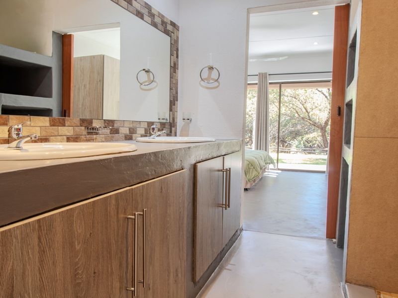 A modern bathroom featuring a double vanity with wood-finish cabinetry, tiled backsplash, and large wall-mounted mirrors, with direct access to the bedroom.