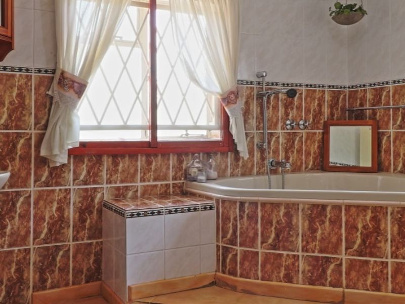 A dated bathroom featuring a built-in bathtub, wall-mounted shower fixtures, and a pedestal basin with traditional brown marble-effect wall tiling.