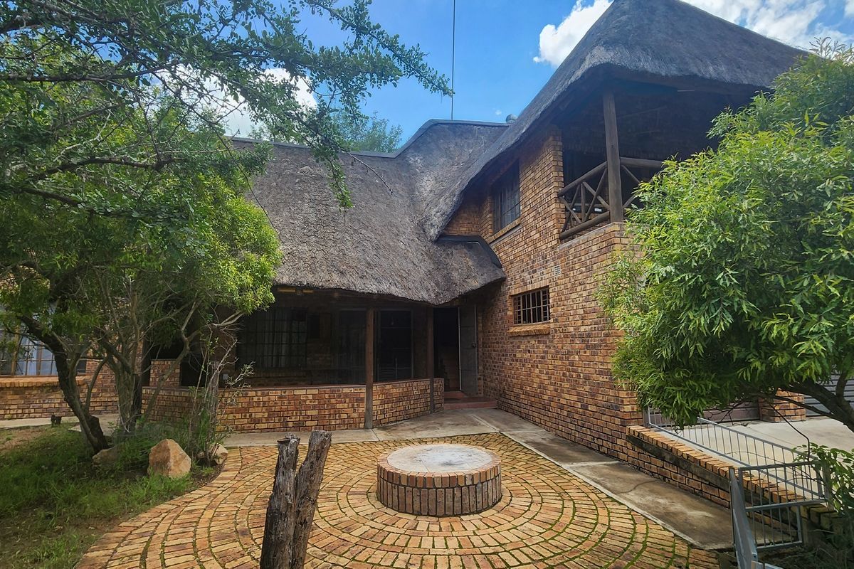 Rear view of a face brick house featuring a traditional thatched roof, a covered veranda, an upper balcony, and a circular brick fire pit area.