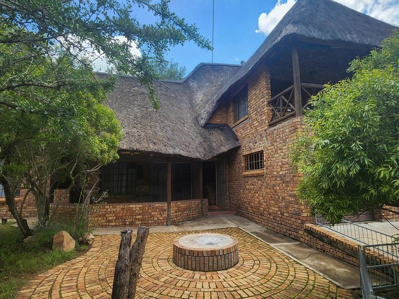 Rear view of a face brick house featuring a traditional thatched roof, a covered veranda, an upper balcony, and a circular brick fire pit area.