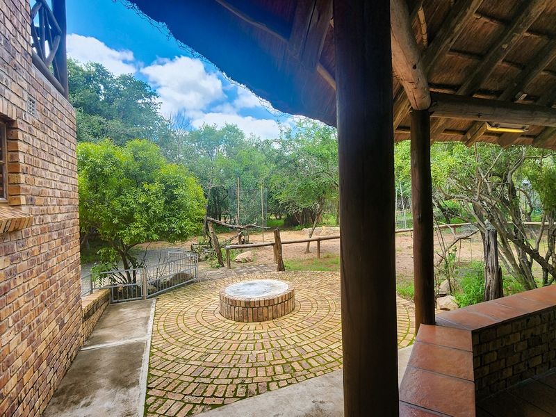 A covered outdoor patio area featuring a thatched roof, timber columns, built-in brick fire pit, and tiled bench seating overlooking the garden.