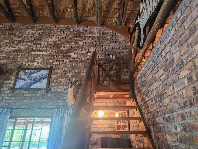 Rustic living area featuring exposed brick walls, a high thatched ceiling with timber rafters, and a wooden staircase leading to an upper level.