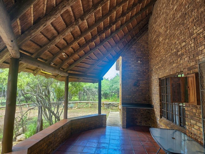 A covered patio area featuring a traditional thatched roof, exposed timber rafters, brick walls, and a built-in braai, overlooking the garden.