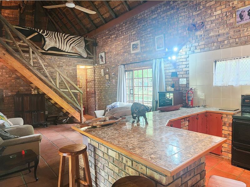 Open-plan living area featuring exposed brick walls, a high thatched ceiling, timber staircase, and a functional kitchen with a tiled island and built-in oven.