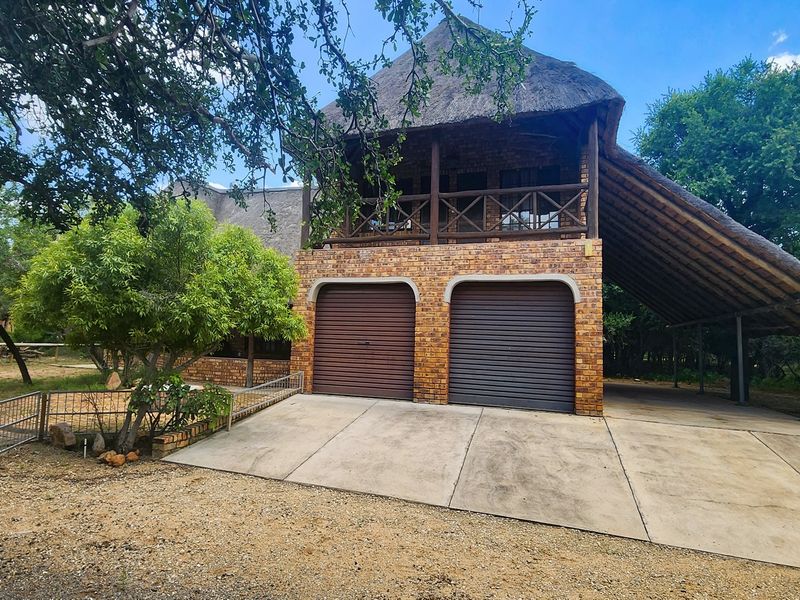 Front view of a double-story brick house featuring a thatched roof, double garage, and an attached carport set in a natural bushveld environment.
