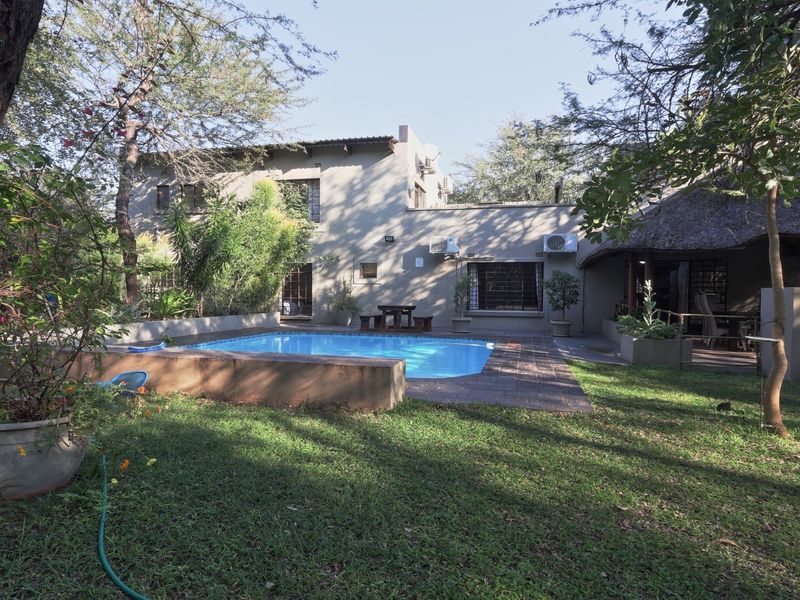 A private backyard featuring a swimming pool, paved patio area, and a thatched roof lapa, surrounded by established trees and garden space.