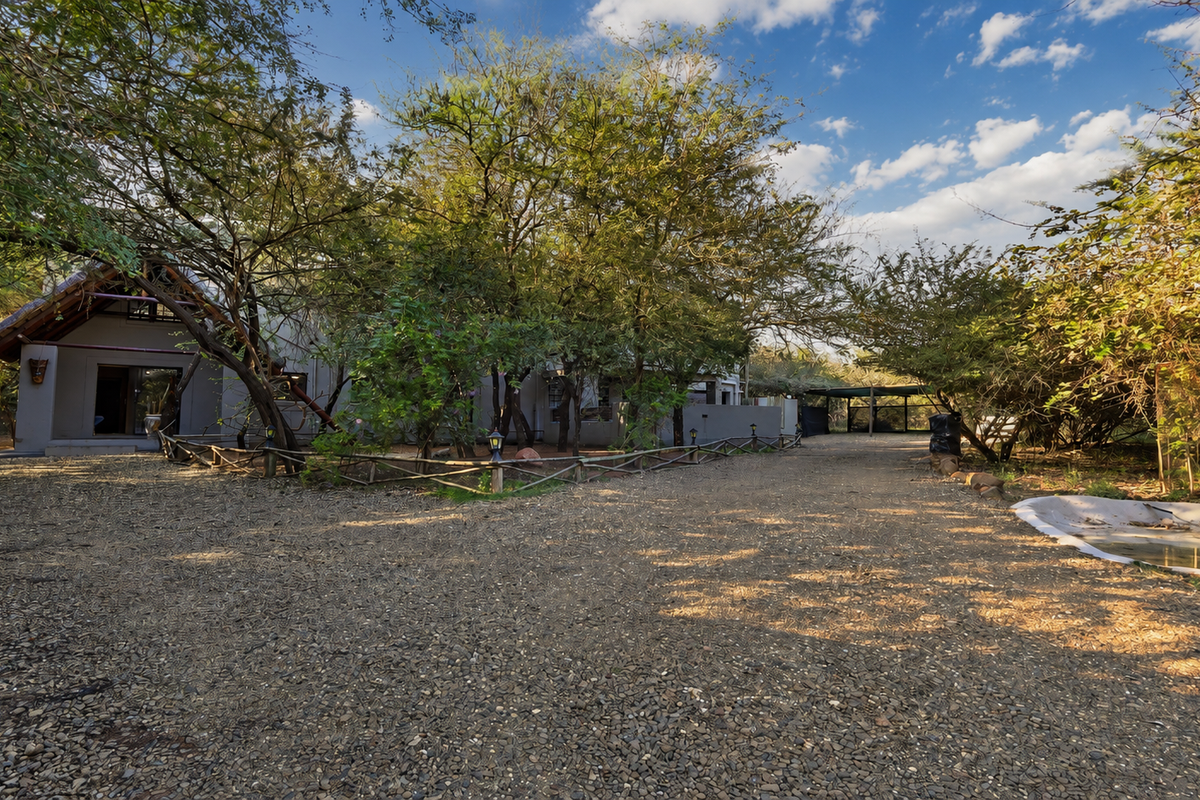 A rustic rear exterior view featuring a gravel driveway, mature trees, timber fencing, and a small water feature set against a natural bushveld backdrop.
