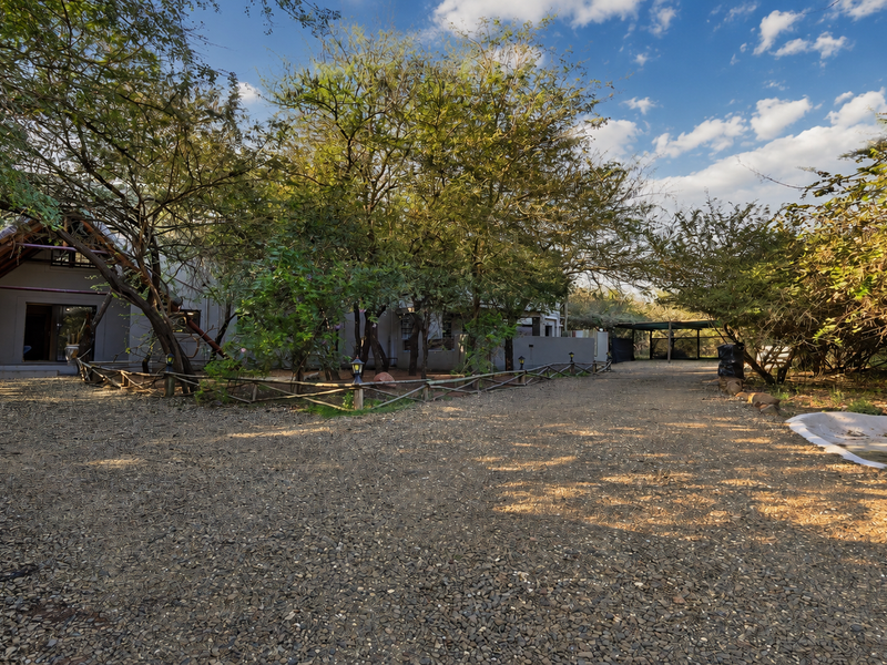 A rustic rear exterior view featuring a gravel driveway, mature trees, timber fencing, and a small water feature set against a natural bushveld backdrop.