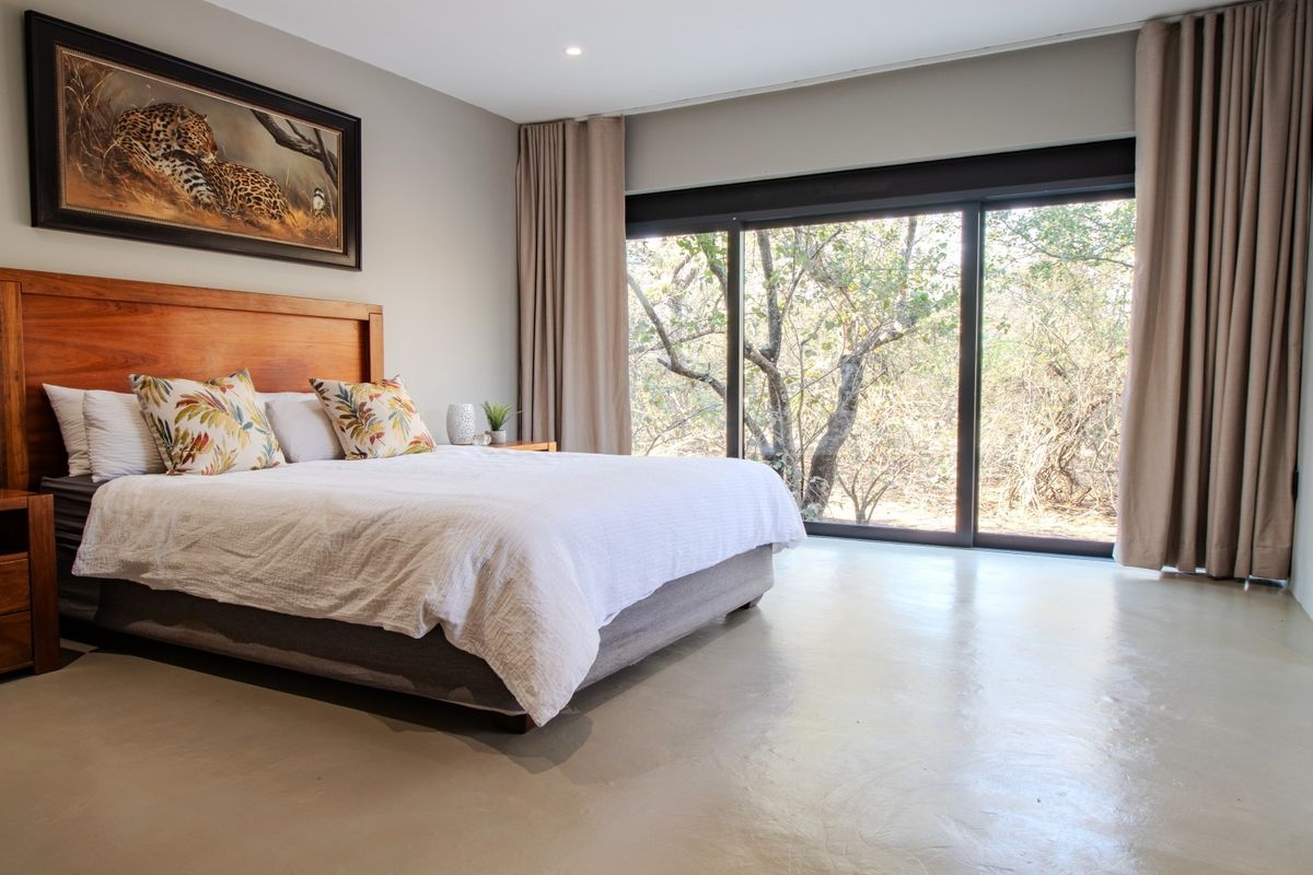 Spacious bedroom featuring polished concrete floors, recessed lighting, and large sliding glass doors providing natural light and views of the surrounding bushveld.