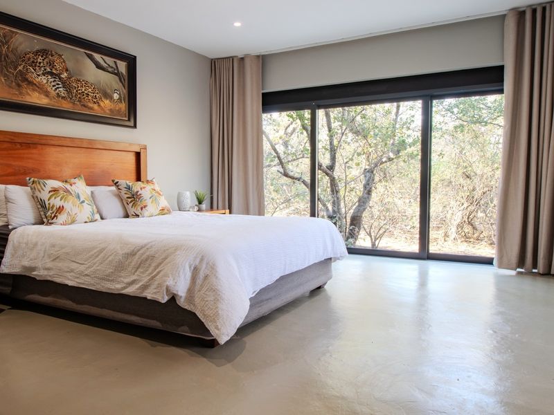 Spacious bedroom featuring polished concrete floors, recessed lighting, and large sliding glass doors providing natural light and views of the surrounding bushveld.