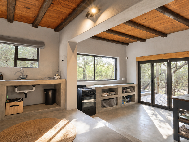 A rustic-modern kitchen featuring exposed timber beams, polished concrete floors, built-in concrete cabinetry, and large sliding doors opening to an outdoor area.