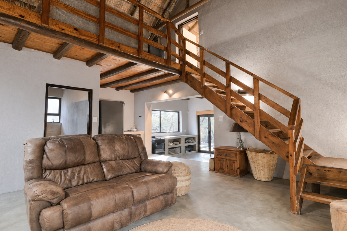 Spacious living area featuring exposed timber beams, a thatched roof, concrete flooring, and a wooden staircase leading to a mezzanine level.
