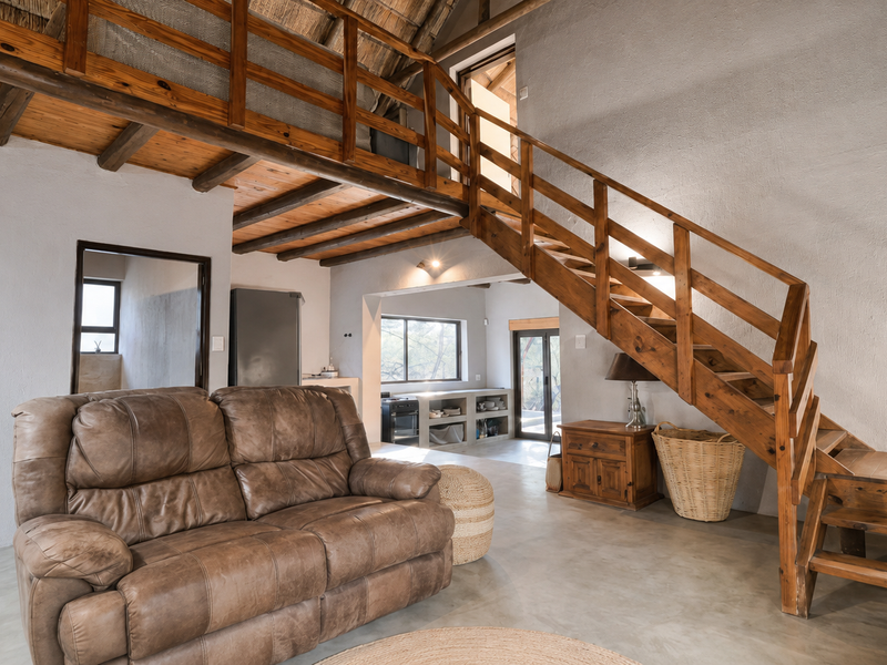 Spacious living area featuring exposed timber beams, a thatched roof, concrete flooring, and a wooden staircase leading to a mezzanine level.