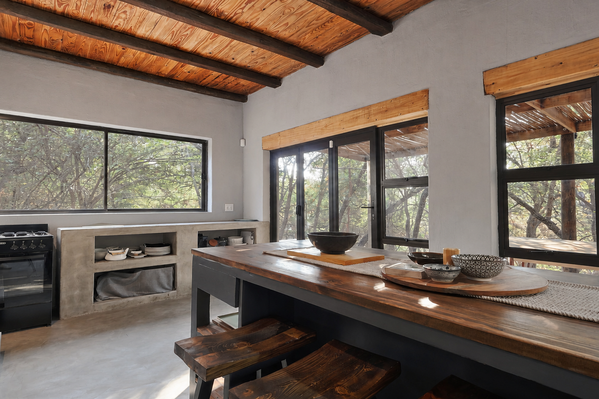 Modern rustic kitchen featuring a concrete island, exposed timber ceiling beams, polished concrete floors, and large black aluminium windows overlooking the surrounding trees.