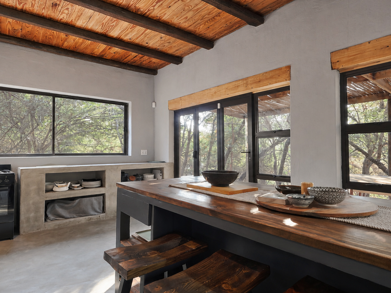 Modern rustic kitchen featuring a concrete island, exposed timber ceiling beams, polished concrete floors, and large black aluminium windows overlooking the surrounding trees.