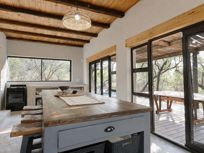 Open plan kitchen and dining area featuring exposed timber beam ceilings, concrete finishes, and large sliding doors opening onto a wooden deck.