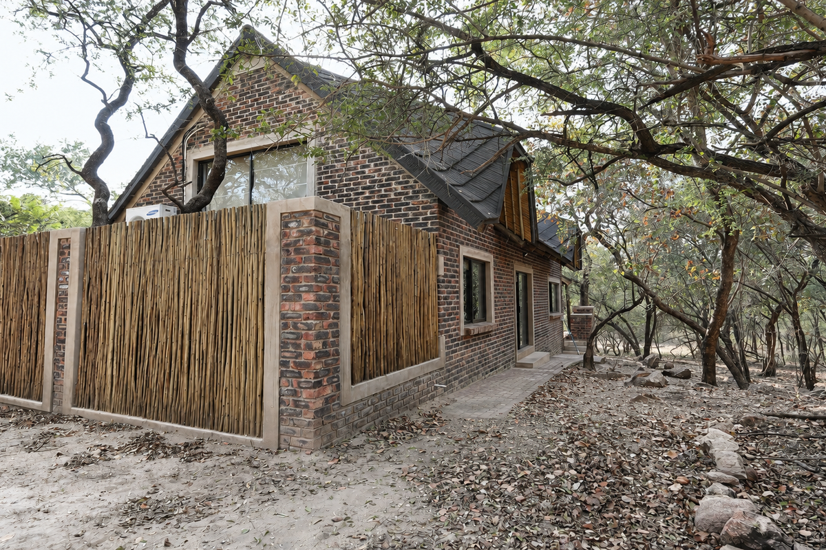 A rear view of a face brick house featuring a bamboo privacy screen, pitched roof, and a paved walkway surrounded by natural woodland.