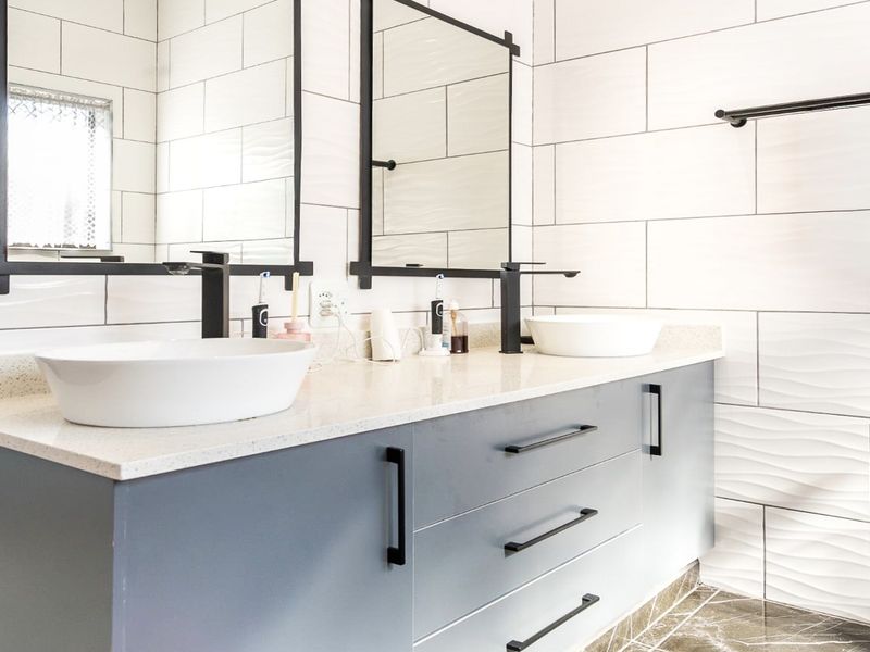 Modern bathroom featuring a double vanity with vessel sinks, matte black fixtures, stone countertop, and contemporary textured wall tiling.