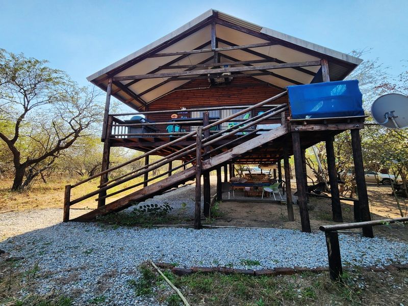 A raised timber cabin featuring a large deck, external staircase, and stilted foundation, set within a natural bushveld environment.