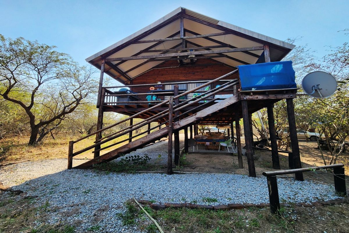 A raised timber cabin featuring a large deck, external staircase, and stilted foundation, set within a natural bushveld environment.