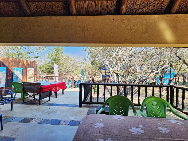 Covered outdoor patio featuring a rustic thatched roof, exposed brick columns, and timber railings, overlooking a natural garden setting.