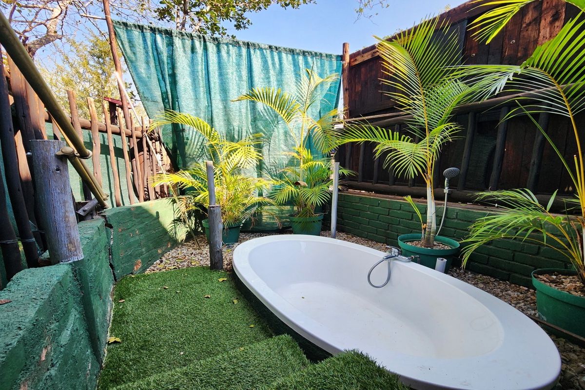 An outdoor bathroom area featuring a freestanding bathtub, handheld shower fixture, and artificial grass set against a rustic brick and timber boundary wall.