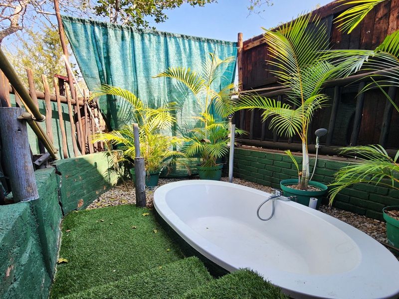 An outdoor bathroom area featuring a freestanding bathtub, handheld shower fixture, and artificial grass set against a rustic brick and timber boundary wall.