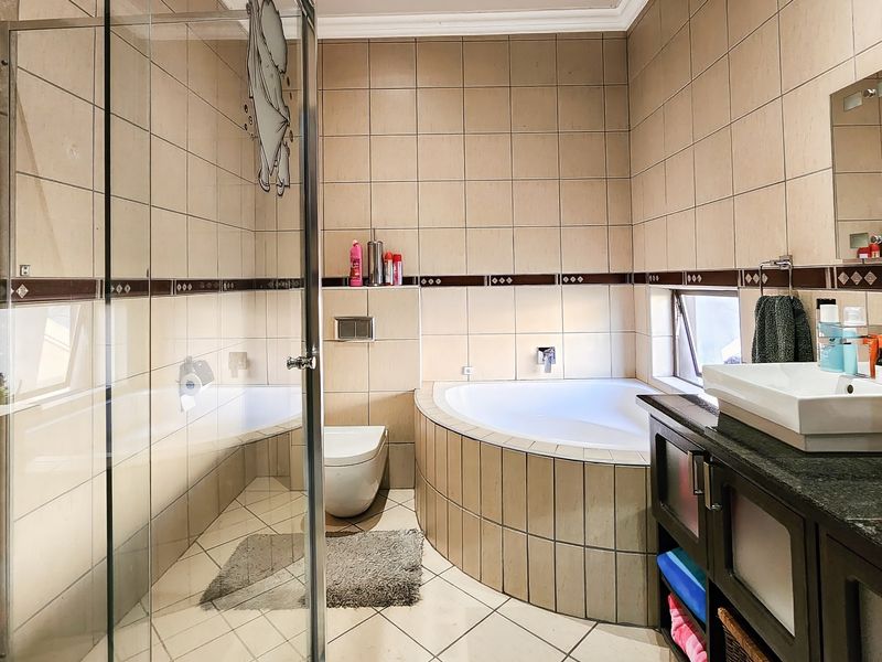 A functional bathroom featuring a corner bathtub, glass-enclosed shower, wall-mounted toilet, and a double vanity with tiled finishes throughout.