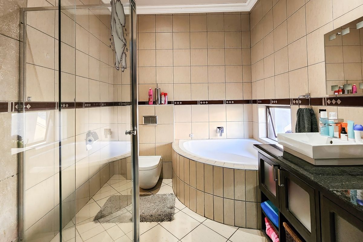 A functional bathroom featuring a corner bathtub, glass-enclosed shower, wall-mounted toilet, and a double vanity with tiled finishes throughout.
