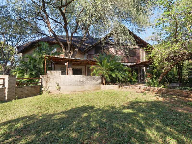Rear view of a multi-story house featuring a thatched roof, brick boundary walls, and a timber pergola set within a mature garden.