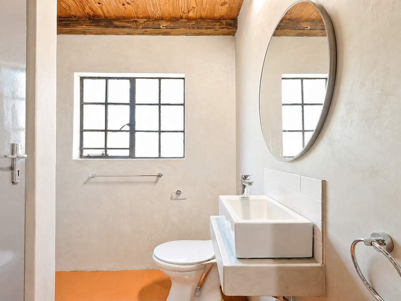 A clean, minimalist bathroom featuring a wall-mounted basin, toilet, and exposed timber ceiling beams, finished with smooth plaster walls and tiled flooring.