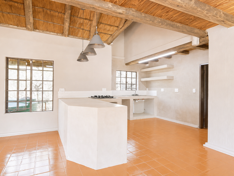 Open-plan kitchen featuring a rustic thatched ceiling, exposed timber beams, built-in concrete island with gas hob, and terracotta-style tiled flooring.