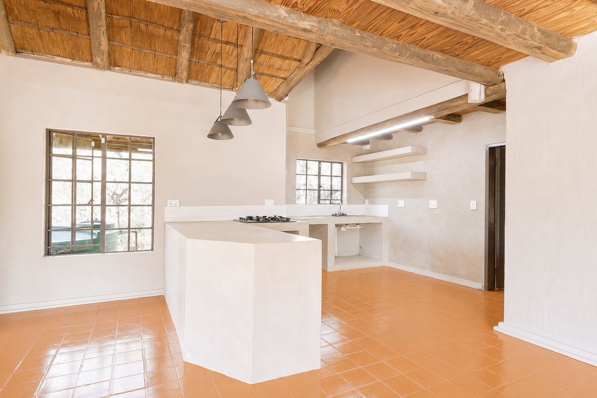 Open-plan kitchen featuring a rustic thatched ceiling, exposed timber beams, built-in concrete island with gas hob, and terracotta-style tiled flooring.