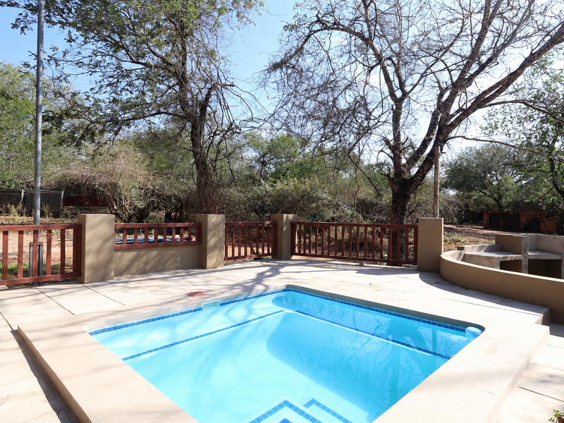 Outdoor pool area featuring a tiled swimming pool, built-in masonry braai, and timber balustrade, set within a natural garden environment.