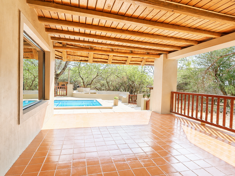 A spacious covered patio featuring exposed timber beam ceilings and terracotta tile flooring, overlooking a private swimming pool and natural garden surroundings.