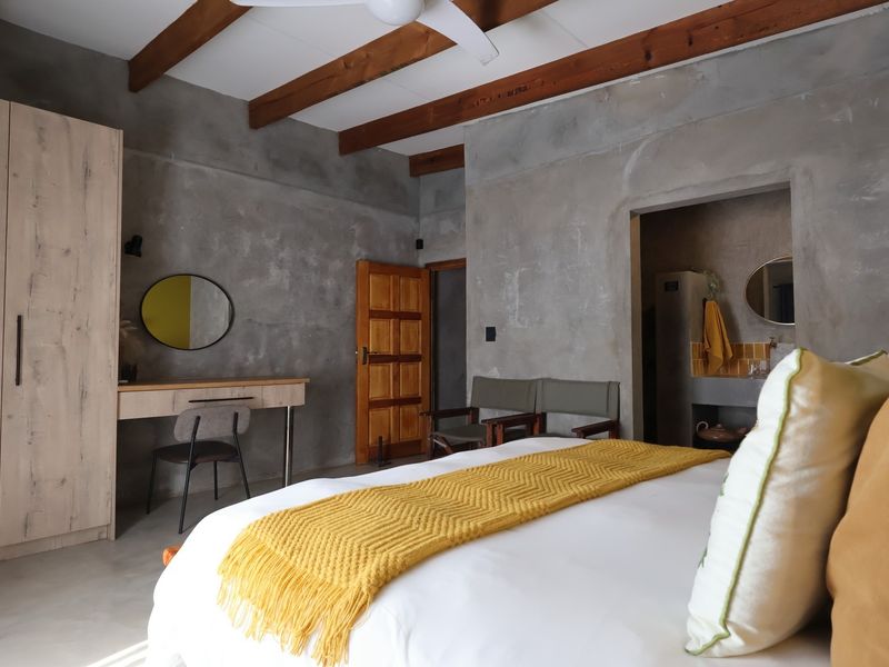 Spacious bedroom featuring exposed timber ceiling beams, industrial-style cement screed walls, polished concrete floors, and a convenient built-in wardrobe and vanity area.