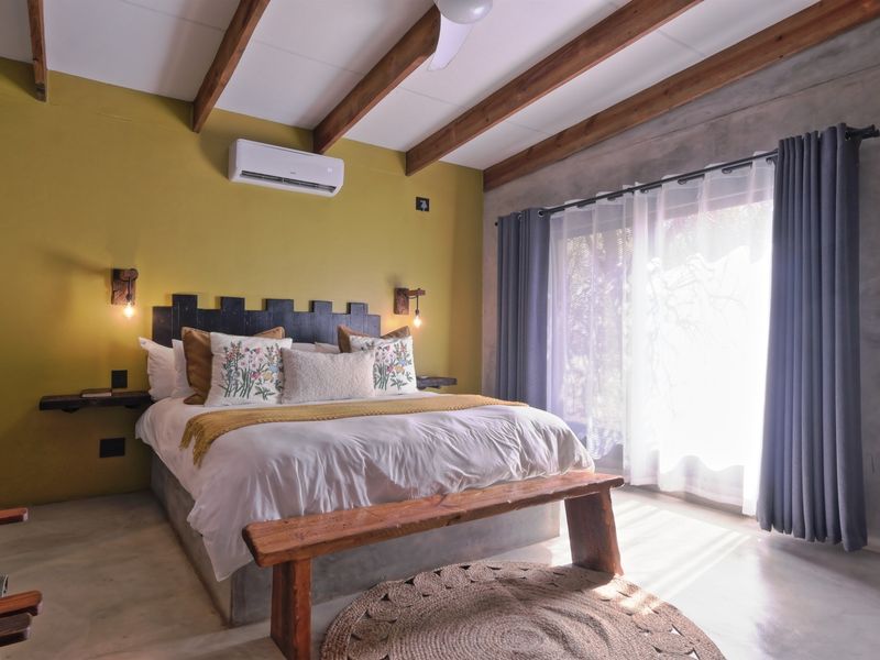 A well-maintained bedroom featuring exposed timber ceiling beams, polished concrete flooring, a built-in concrete bed base, and a split-system air conditioner.