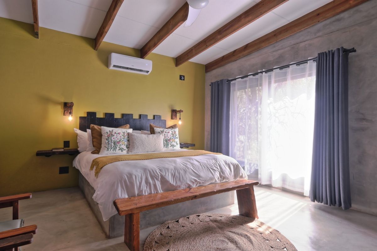 A well-maintained bedroom featuring exposed timber ceiling beams, polished concrete flooring, a built-in concrete bed base, and a split-system air conditioner.