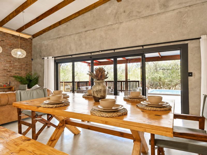Spacious open-plan living and dining area featuring exposed timber beams, a brick feature wall, polished concrete floors, and seamless access to the outdoor patio.