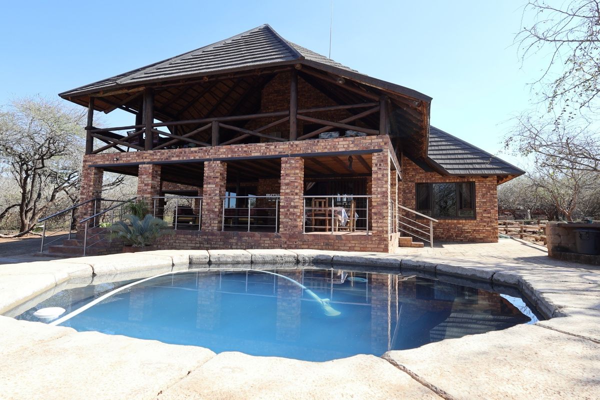 A spacious outdoor pool area featuring a swimming pool with stone coping, set against a rustic brick house with a large covered patio.