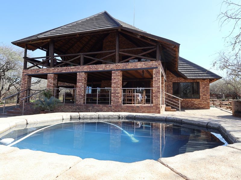 A spacious outdoor pool area featuring a swimming pool with stone coping, set against a rustic brick house with a large covered patio.