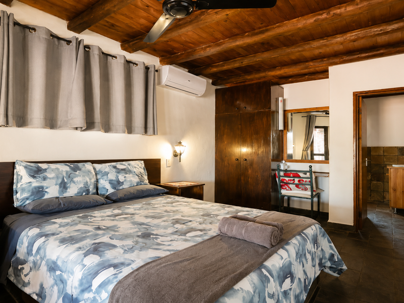 A bedroom featuring exposed timber ceiling beams, built-in wooden cabinetry, wall-mounted air conditioning, and tiled flooring, with an adjoining bathroom visible.