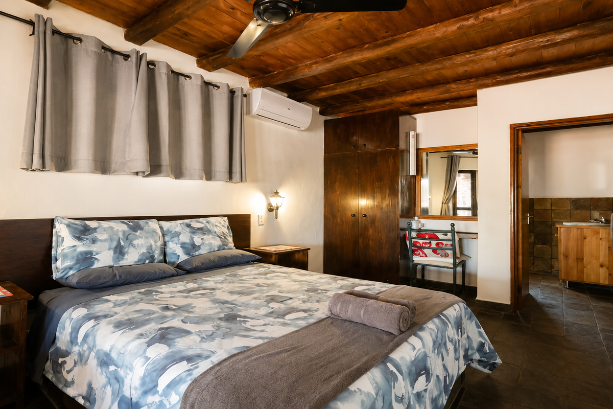 A bedroom featuring exposed timber ceiling beams, built-in wooden cabinetry, wall-mounted air conditioning, and tiled flooring, with an adjoining bathroom visible.