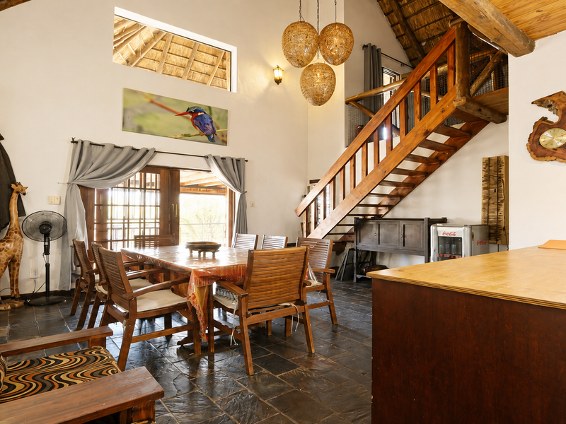 Open plan living and dining area featuring high vaulted thatched ceilings, exposed timber beams, a wooden staircase, and slate tile flooring.