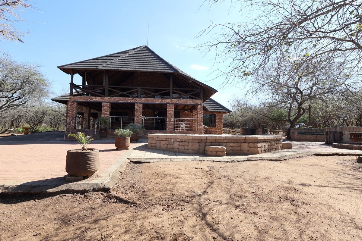 Rear view of a rustic brick house featuring a covered patio, timber balcony, and a stone-walled terrace set within a natural bushveld landscape.