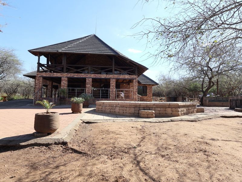 Rear view of a rustic brick house featuring a covered patio, timber balcony, and a stone-walled terrace set within a natural bushveld landscape.