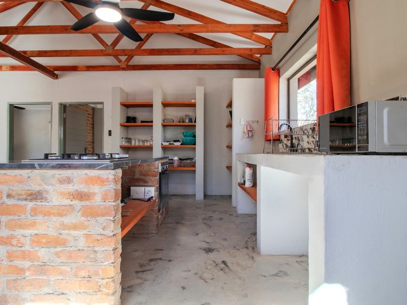 Rustic kitchen featuring exposed timber roof trusses, a brick island with a gas hob, concrete countertops, and open built-in shelving.